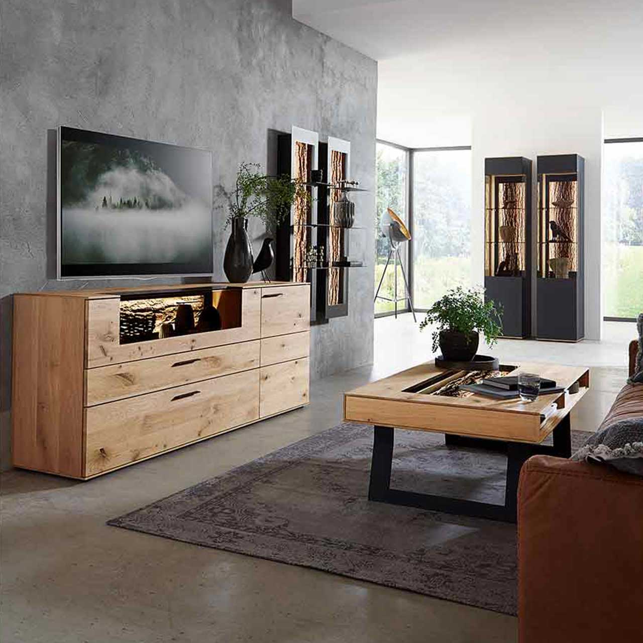 Wohnzimmer, in dem rechts ein Sideboard aus hellem Holz steht und rechts auf einem Teppich ein Couchtisch auf Kufen vor einem Sofa. Im Hintergrund sind zwei Hängeelemente und zwei Standvitrinen zu sehen.