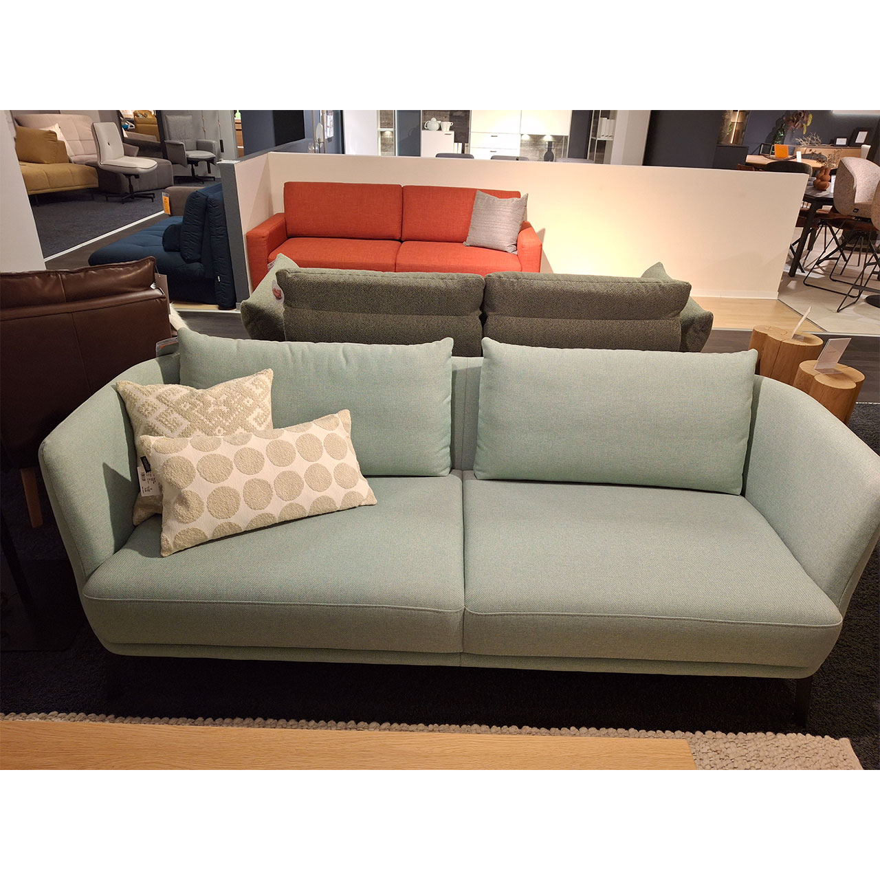 Sofa mit drei Sitzplätzen und zwei Rückenkissen mit schmalen Armlehnen. Im Hintergrund sind weitere Sofas in einer Ausstellung zu sehen.