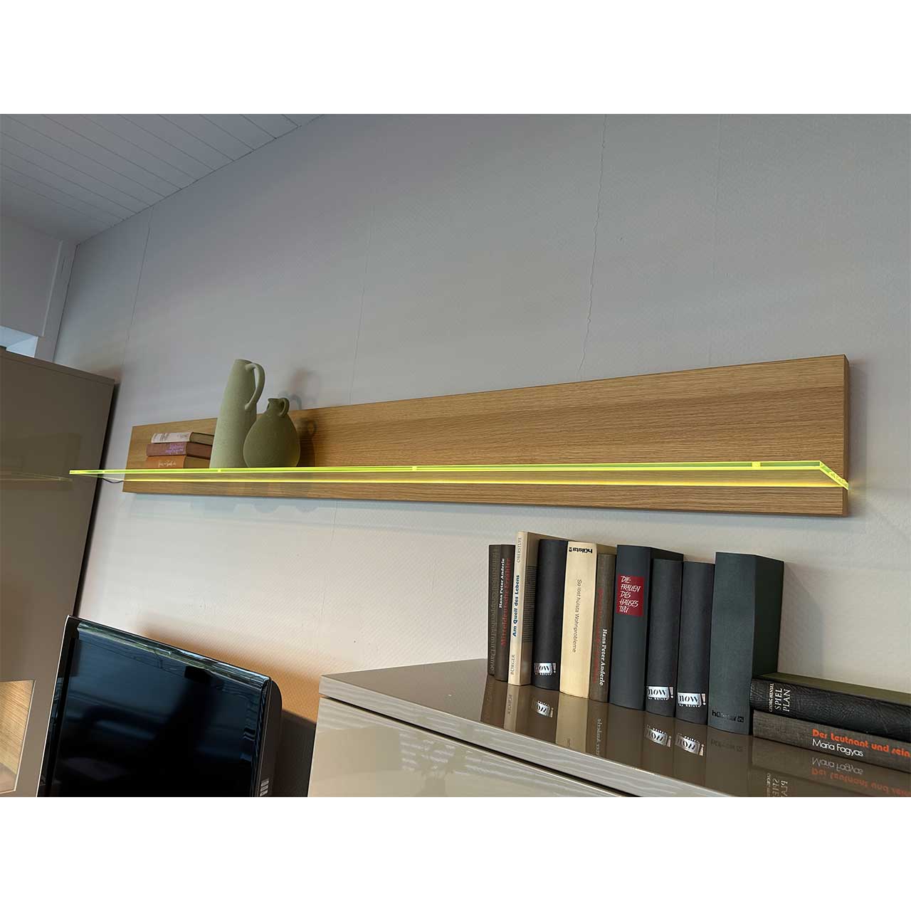 Das Wandbord aus hellem Holz hat eine beleuchtete gläserne Ablagefläche.