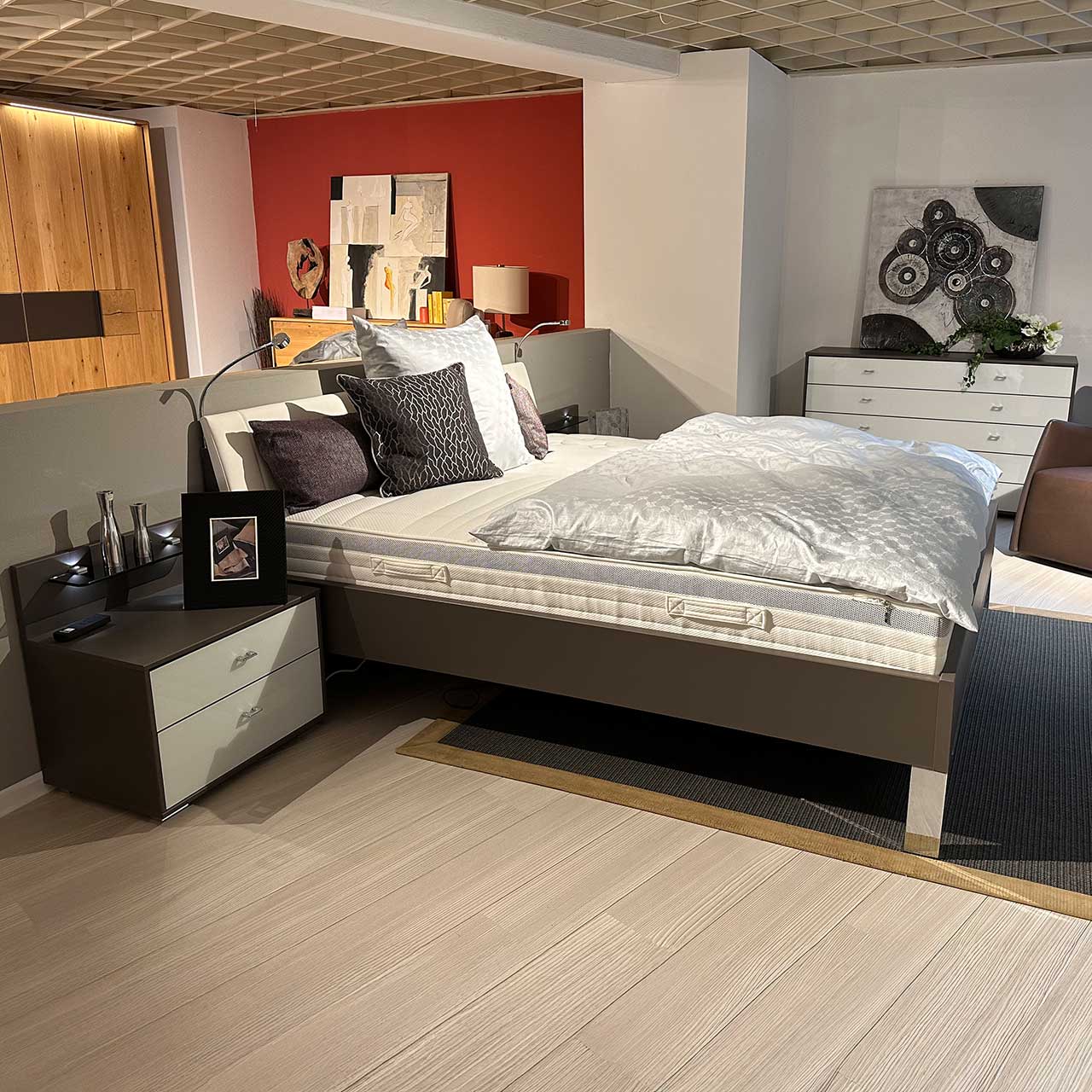 Bett mit dunkelgrauem Bettrahmen und weißem Kopfteil auf chromfarbigen Füßen, daneben rechts und links je eine Nachtkommode im gleichen Look (Dunkelgrauer Korpus, weiße Schubladenfronten).