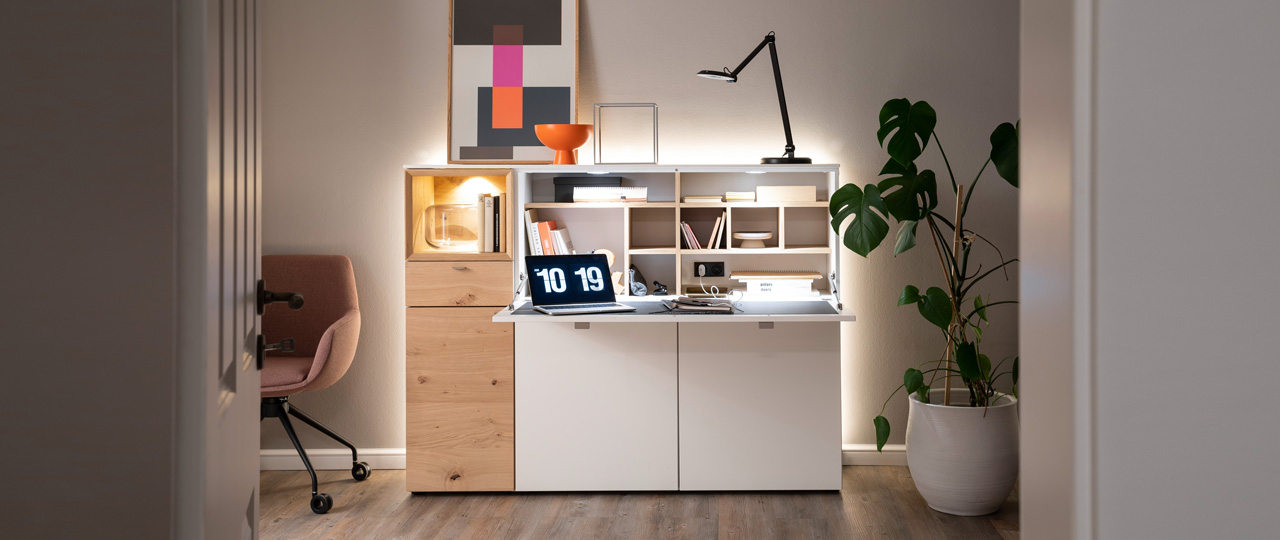 MH_banner_marken_sw Ambientebild der Marke schöner Wohnen mit Fokus auf ein weißes Highboard mit einseitigem Naturholzakzent und Sekretärfunktion.