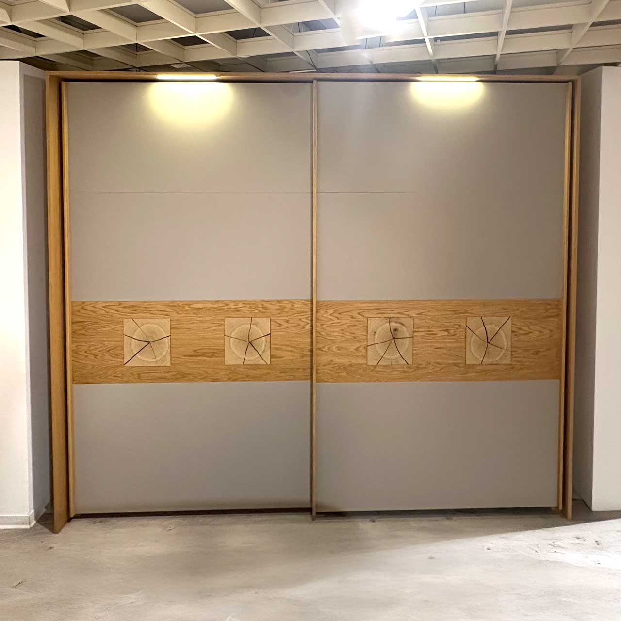 Schöner Wohnen - Schiebetürenschrank Multiline Der Außenkorpus ist aus Holz, die zwei grauen Türen tragen als Zierde ein Holzband, auf dem pro Tür zwei quadratische Hirnholzapplikationen eingearbeitet sind. Oben am Rahmen findet sich je Tür eine Lichtquelle, die den Schrank beleuchtet.
