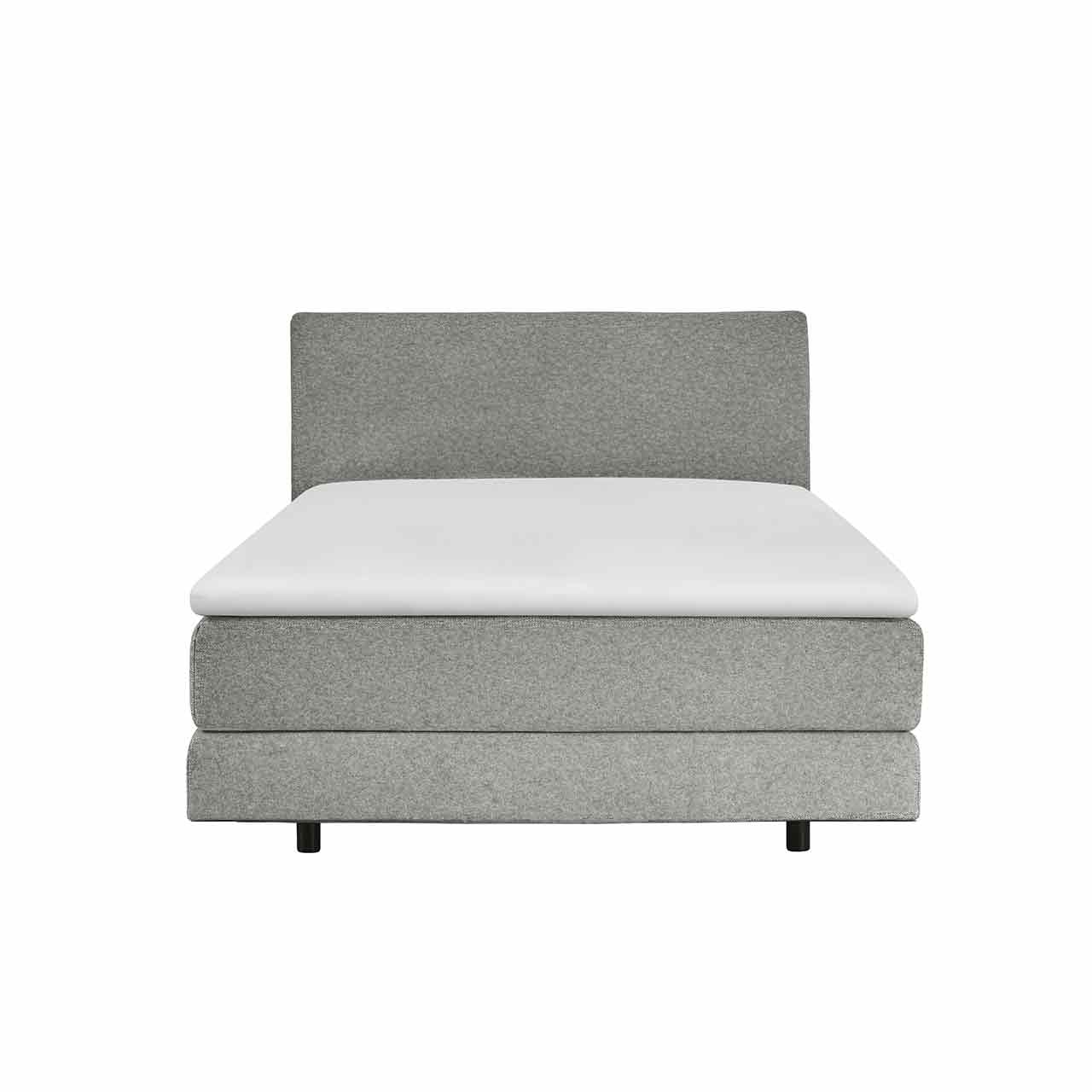 Schmales Boxspringbett in hellgrauen Stoff mit einer Box unten und mit Möbelstoff bezogener Matratze darüber. Darauf liegt ein weißer Topper. Das Kopfteil ist glatt mit dem gleichen Möbelstoff bezogen.
