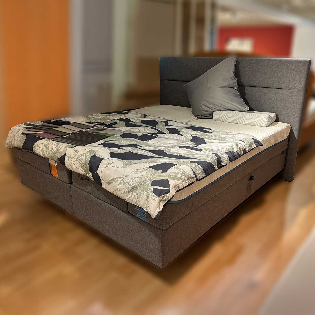 Tempur Boxspringbett Texture - Grau Boxspringbett aus grauem Stoff mit Kopfteil, in dem quer eine Ziernaht eingearbeitet ist.