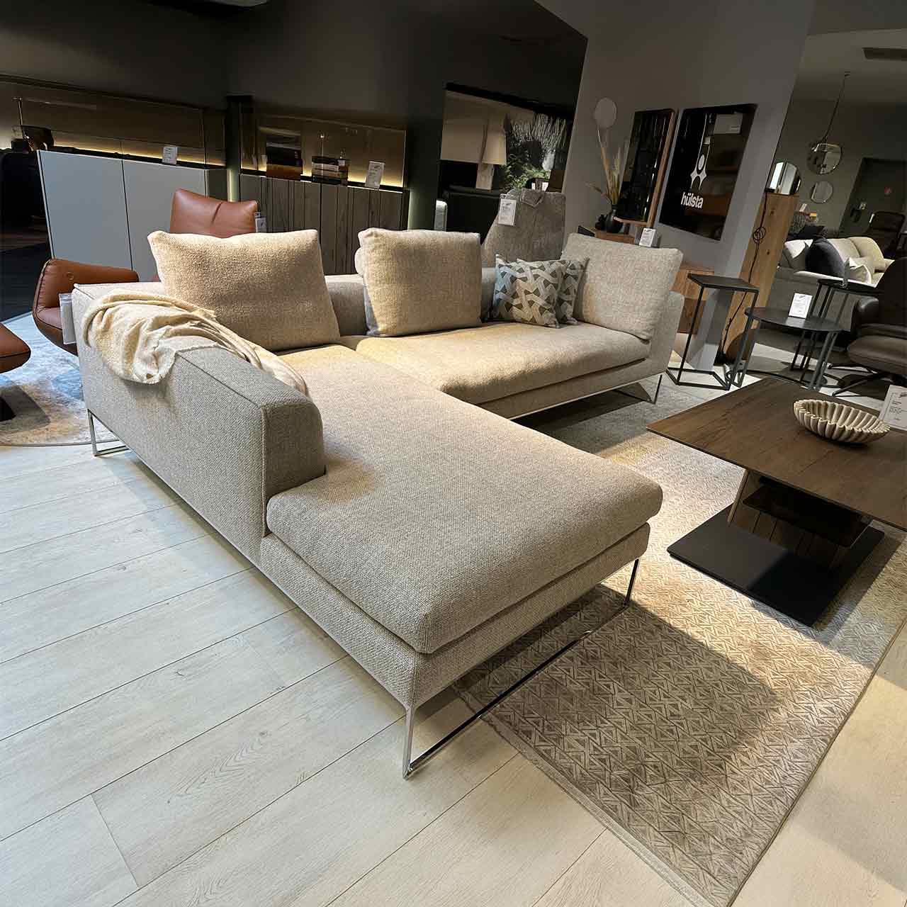 Modernes Ecksofa aus hellem Stoff auf glänzenden Kufen. Links Ottomane, rechts Sitzkombination. Darauf drei Kissen mit Rolle dahinter.