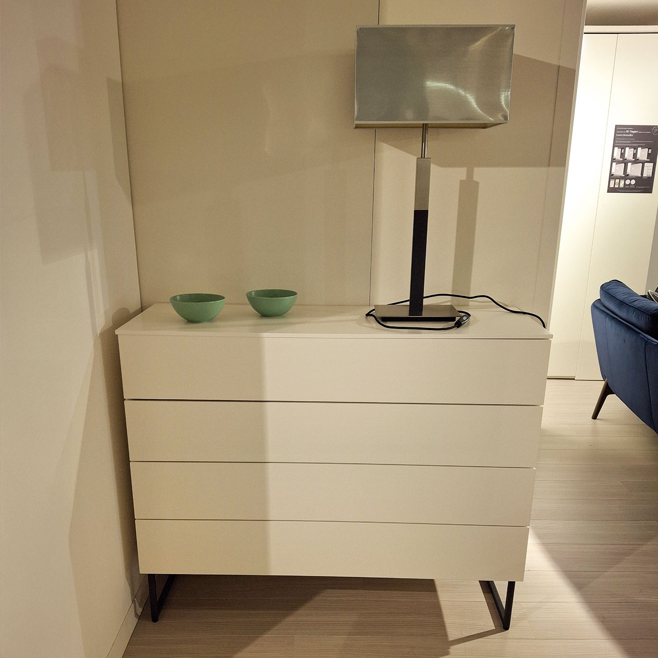 Weiße Kommode mit vier Schubladen und schwarzen Metallkufen. Auf der Kommode steht eine Tischlampe rechts und zwei grüne Schalen.