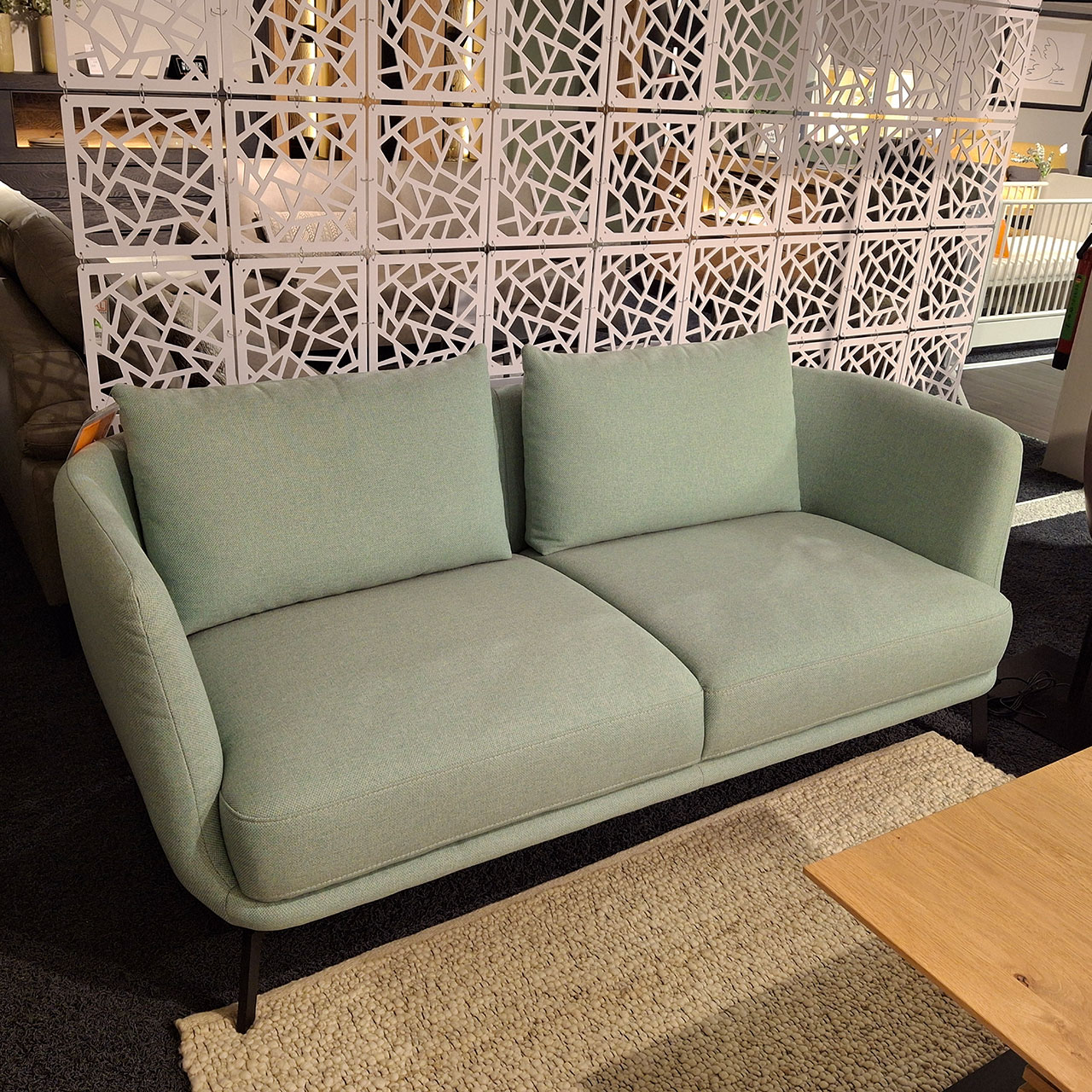 Sofa mit 2,5 Sitzplätzen und zwei Rückenkissen mit schmalen Armlehnen. Im Hintergrund sind weitere Sofas in einer Ausstellung zu sehen.
