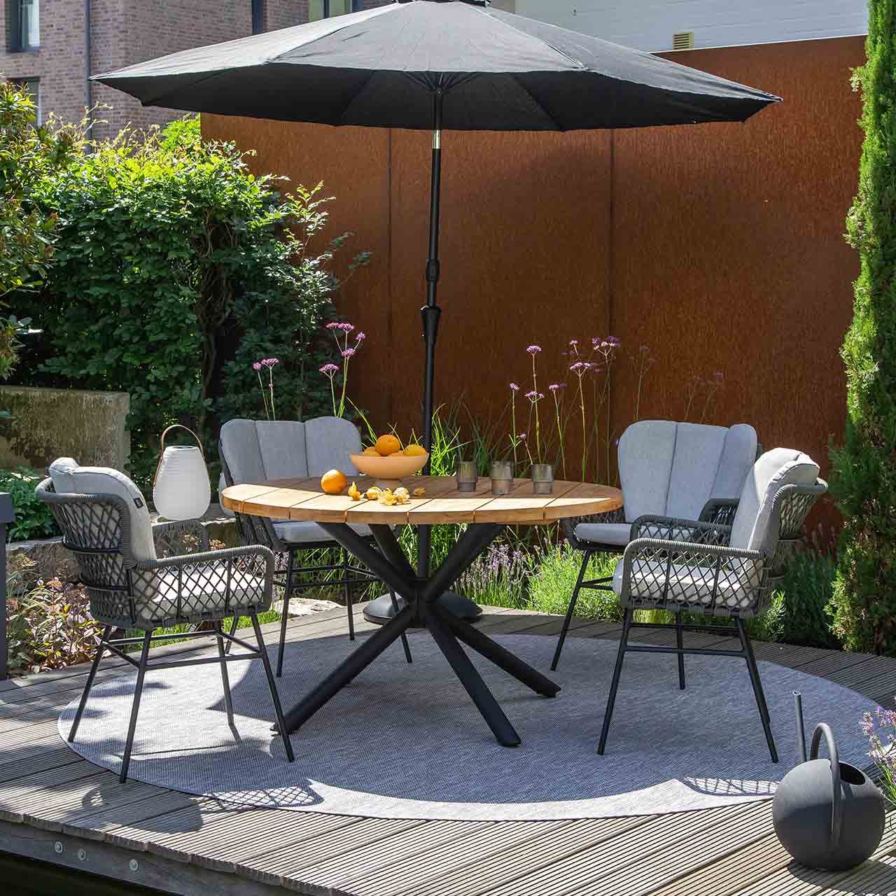 Runder Gartentisch SABA mit Teakholz Platte und Sternförmiges schwarzes Untergestell. Beistehend sind vier Bennett Geflecht Stühle inkl. Kissen.