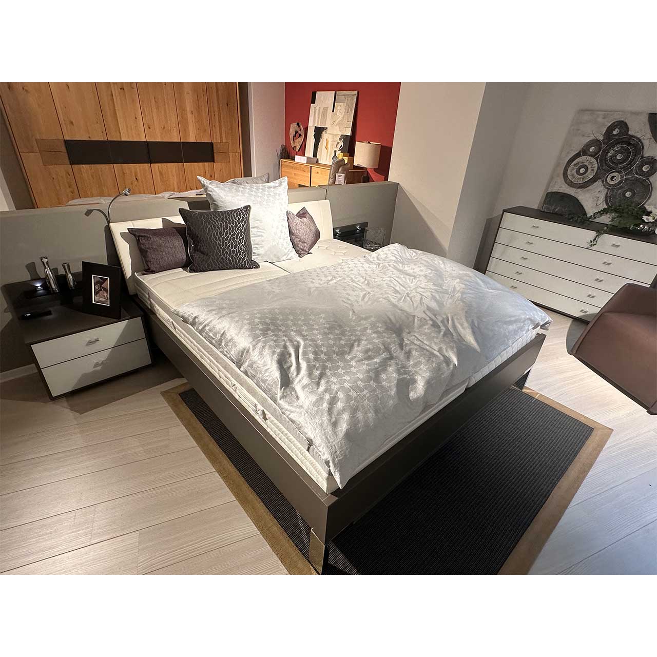 Bett mit dunkelgrauem Bettrahmen und weißem Kopfteil auf chromfarbigen Füßen, daneben rechts und links je eine Nachtkommode im gleichen Look (Dunkelgrauer Korpus, weiße Schubladenfronten).