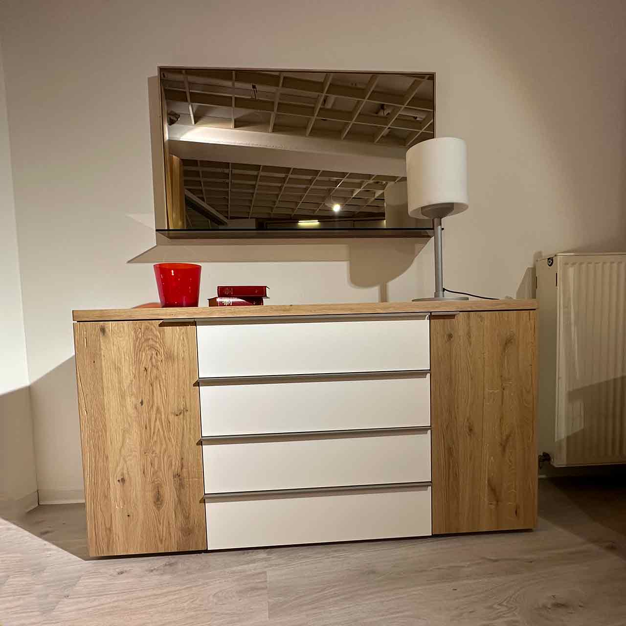 Kommode aus hellem Holz. Rechts und links Holztüren, in der Mitte vier Schubladen mit weißer Front. Darüber ein Spiegel.