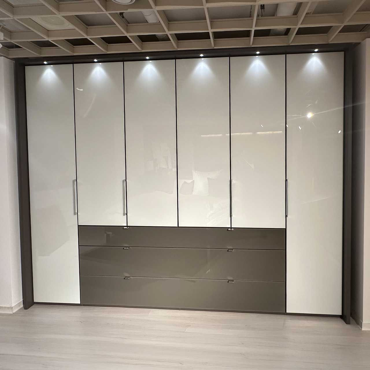 Glänzend weißer Kleiderschrank mit dunkelgrauem Passepartout, in das pro Element eine Leuchte eingebaut ist. Drei dunkelgraue Außen-Schubladen sind in der Mitte eingebaut, d.h. die vier mittleren Türen sind um die Gesamthöhe der Schubladen verkürzt.