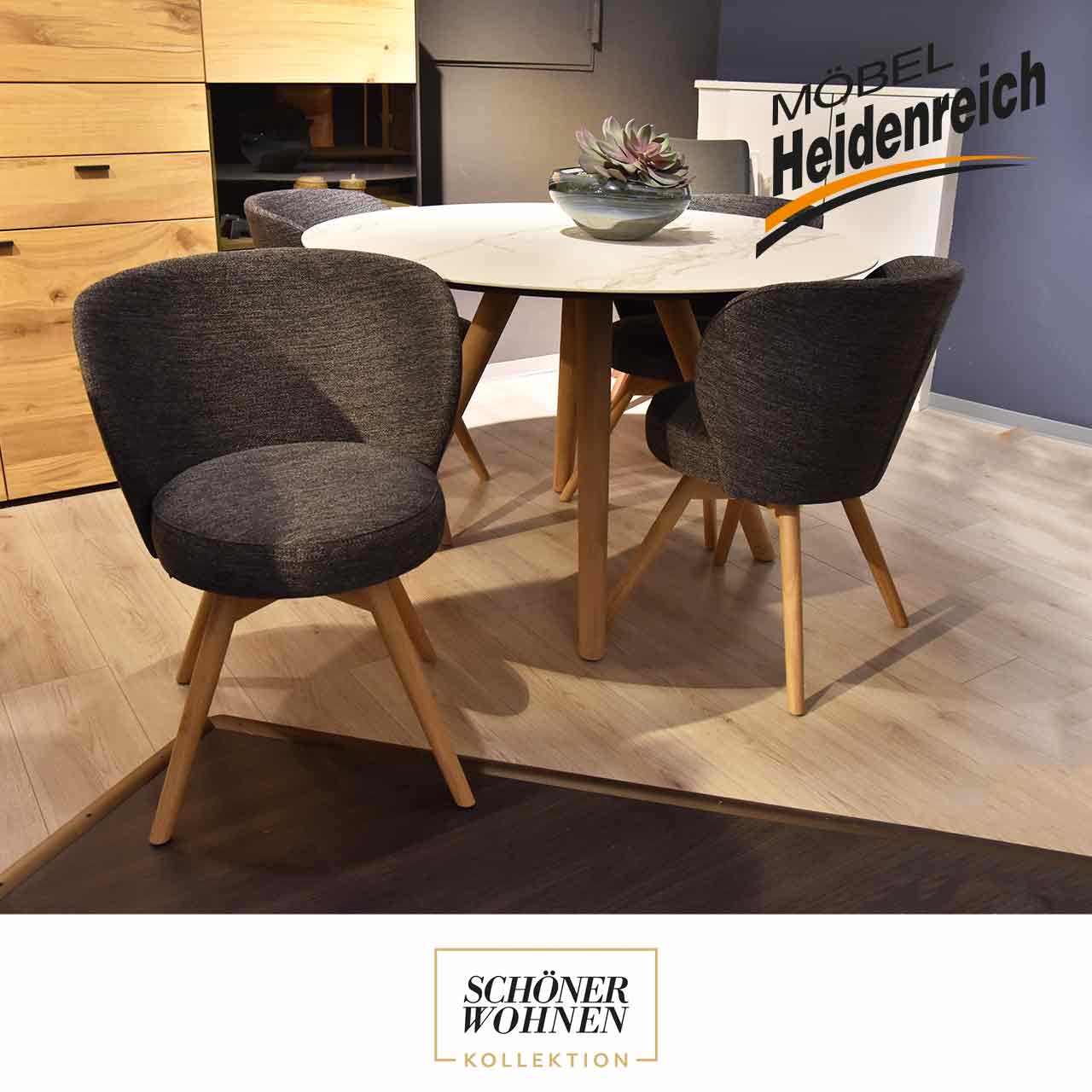Vier Stühle an einem runden Tisch. Ein Stuhl ist nach vorne gedreht. Die Stühle haben einen grauen Bezug und vier helle Holzbeine.