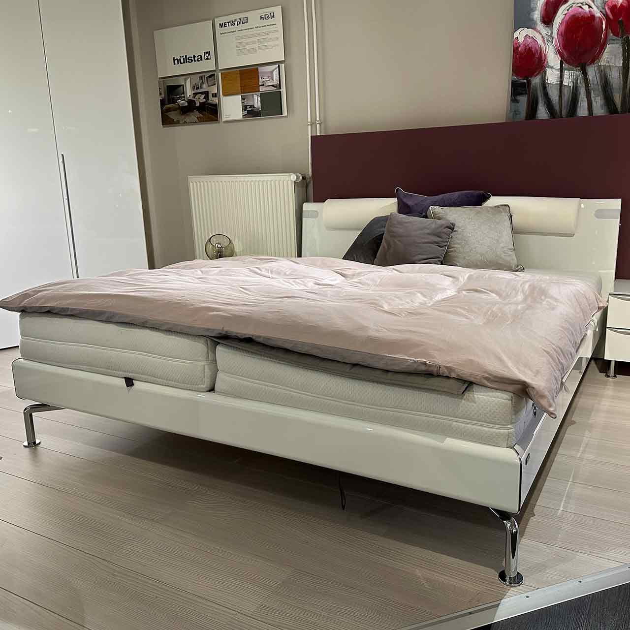 Weißes Bett mit Chrom hochglänzenden Füßen. Kopfteil mit eingearbeiteten Kissen und Bettleseleuchten.