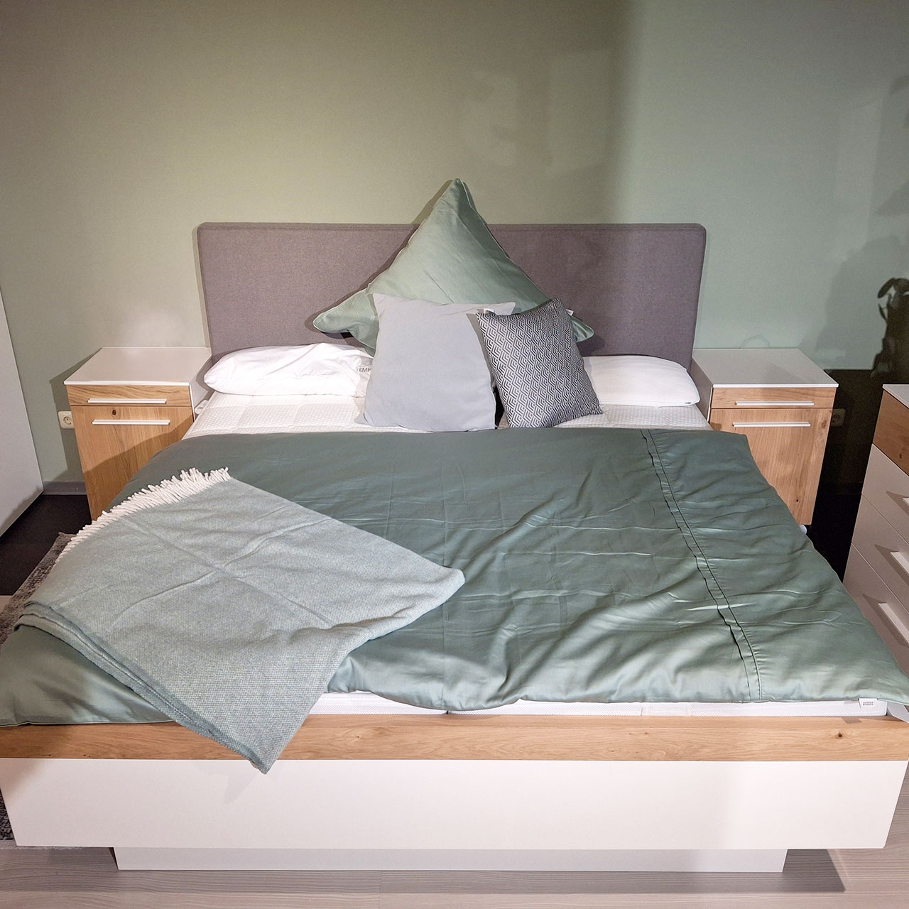Breites Bett mit weißem Rahmen und in Balkeneiche abgesetztem Handlauf. Neben dem Bett stehen zwei passende Konsolen mit Fronten in Balkeneiche und weißen Griffen.