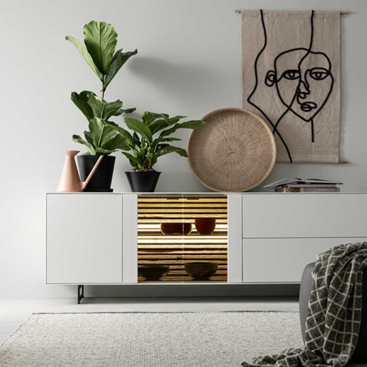 MH_Wohnzimmer_Sideboards_Quadratbild Ambientebild mit modernem Sideboard in Weiß mit Einsatz aus Glas und Beleuchtung, sowie Dekoelementen Pflanzen, Wandbild mit Gesicht und Dekoteller.