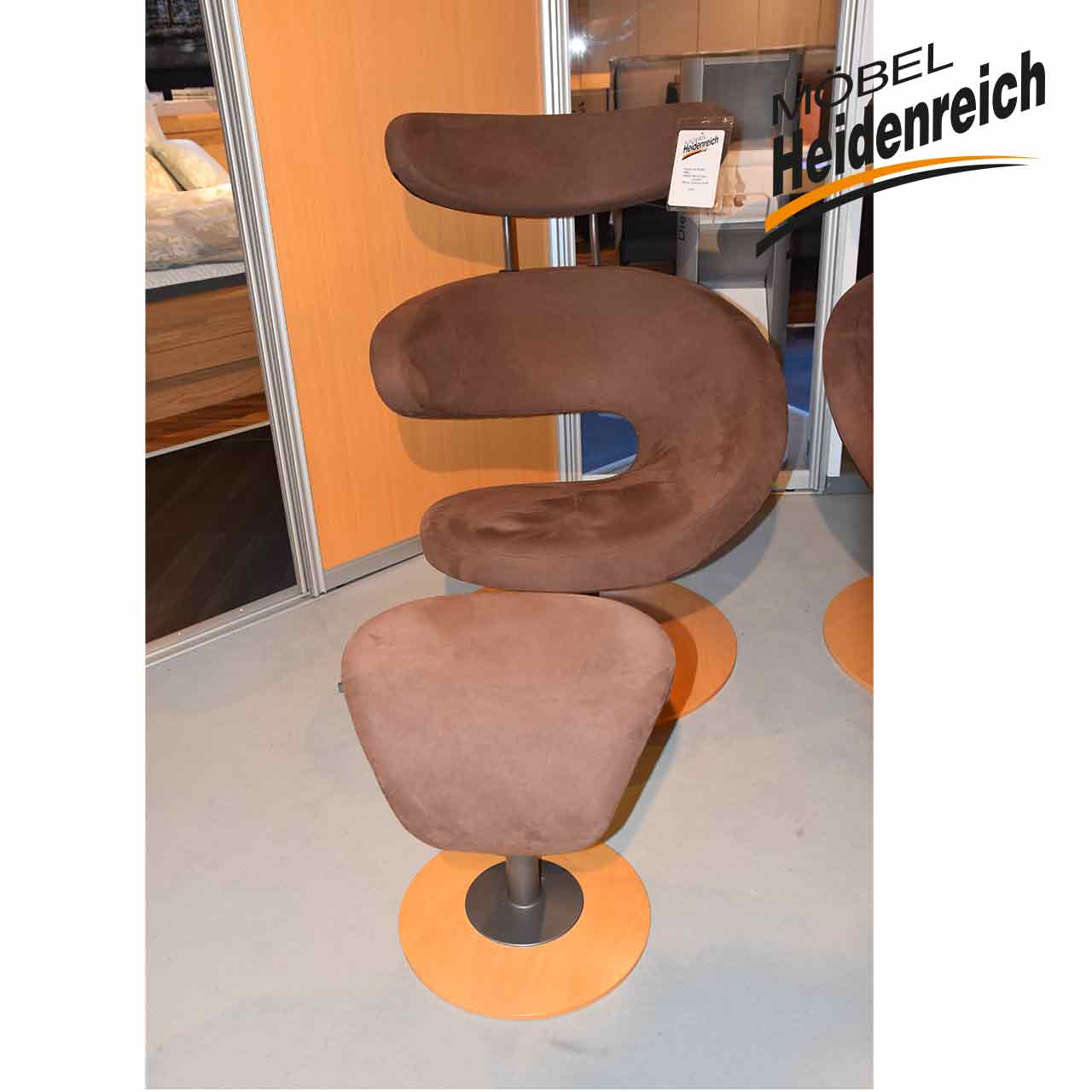 Ausgefallener brauner Sessel, dessen Sitzschale nach rechts geschwungen ist wie bei einer geschälten Orangenschale. Er steht auf einer runden Buchen-Platte.