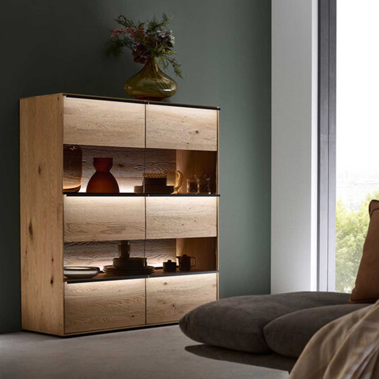 MH_Wohnzimmer_Vitrinen_Quadratbild Ambientebild Vitrinen. Modernes Highboard aus Holz mit Glaselementen. Darauf eine stilvoll, moderne Designerblumenvase.