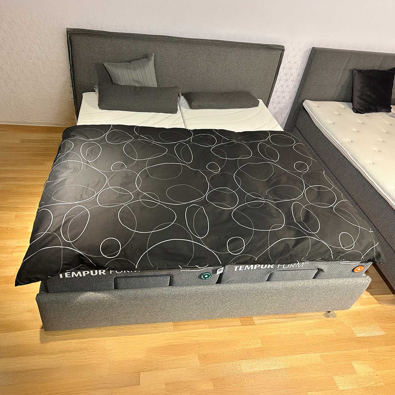 Tempur Boxspringbett Frame Grau im Möbelhaus Das Boxspringbett ist mit glattem grauem Möbelstoff bezogen. Das Kopfteil ist ebenfalls glatt, einzige Zierde die umlaufende Naht.