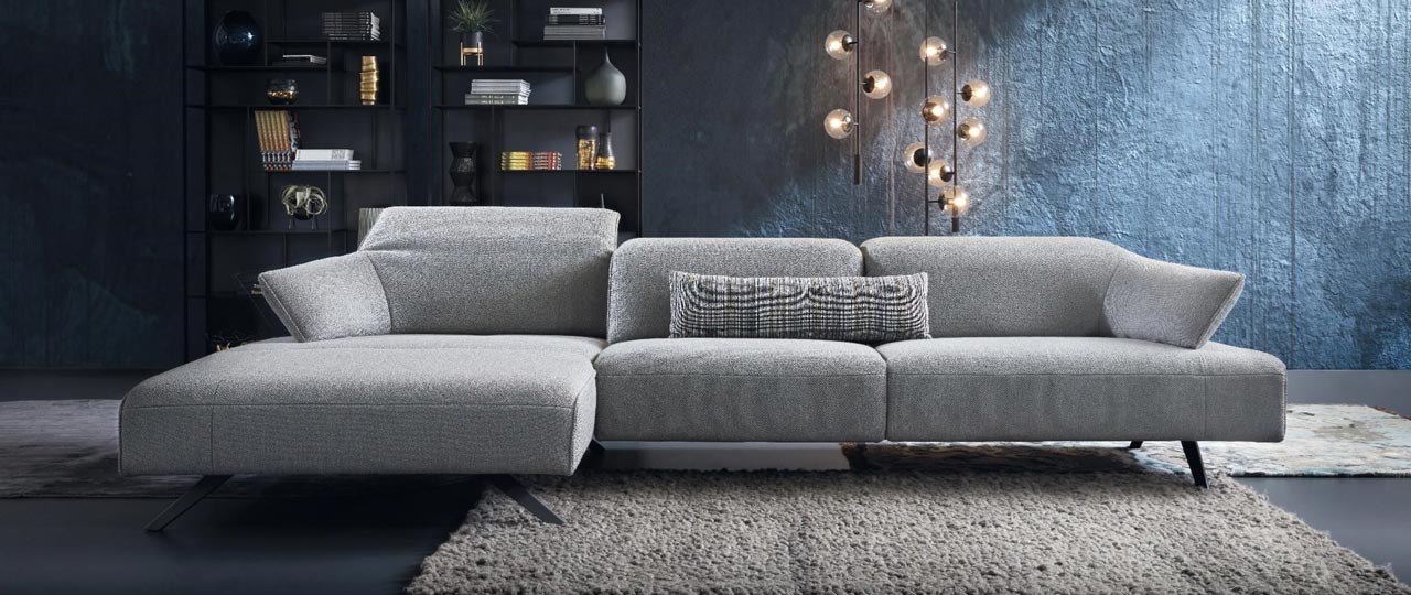 MH_banner_marken_himolla Ambientebild mit Fokus auf ein Sofa der Marke himolla in hellem grau aus Stoff. Im Hintergrund ein modernes schwarzes Regal mit Dekoelementen.