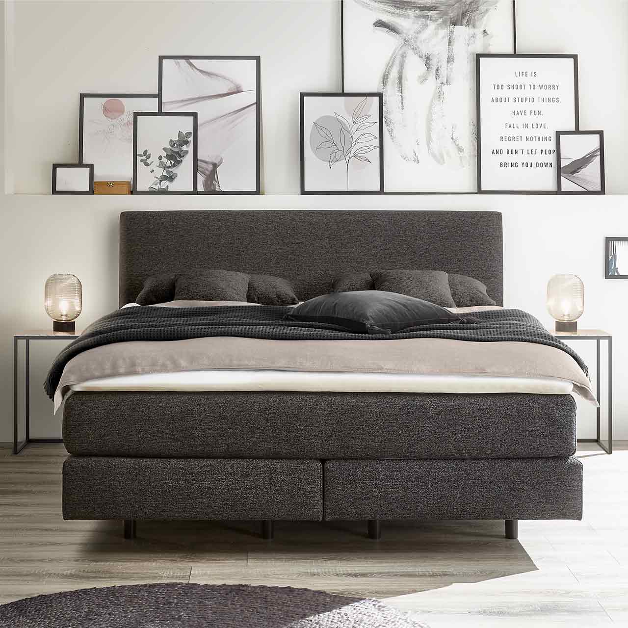Frontalansicht des gleichen Bettes als Milieuaufnahme in einem Schlafzimmer mit viel Dekoration, Decken und Kissen.