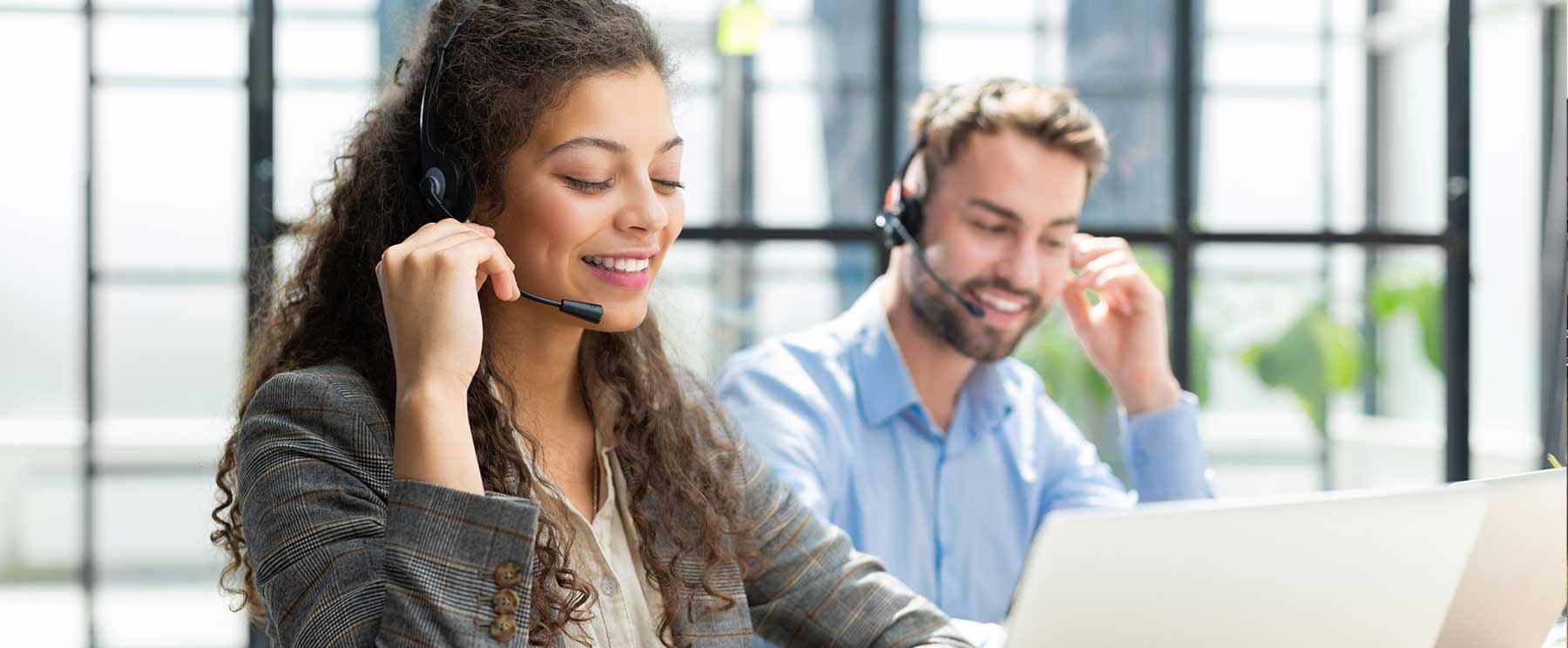 Eine Frau und ein Mann sitzen mit Headsets vor einem Computer und schauen dabei konzentriert aus.