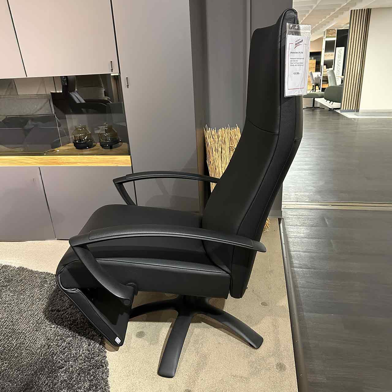 Relaxsessel aus schwarzem glatten Leder mit hohem Kopfteil und eingeklapptem Fußteil von der Seite fotografiert, schwarzer Sternfuß ist sichtbar.