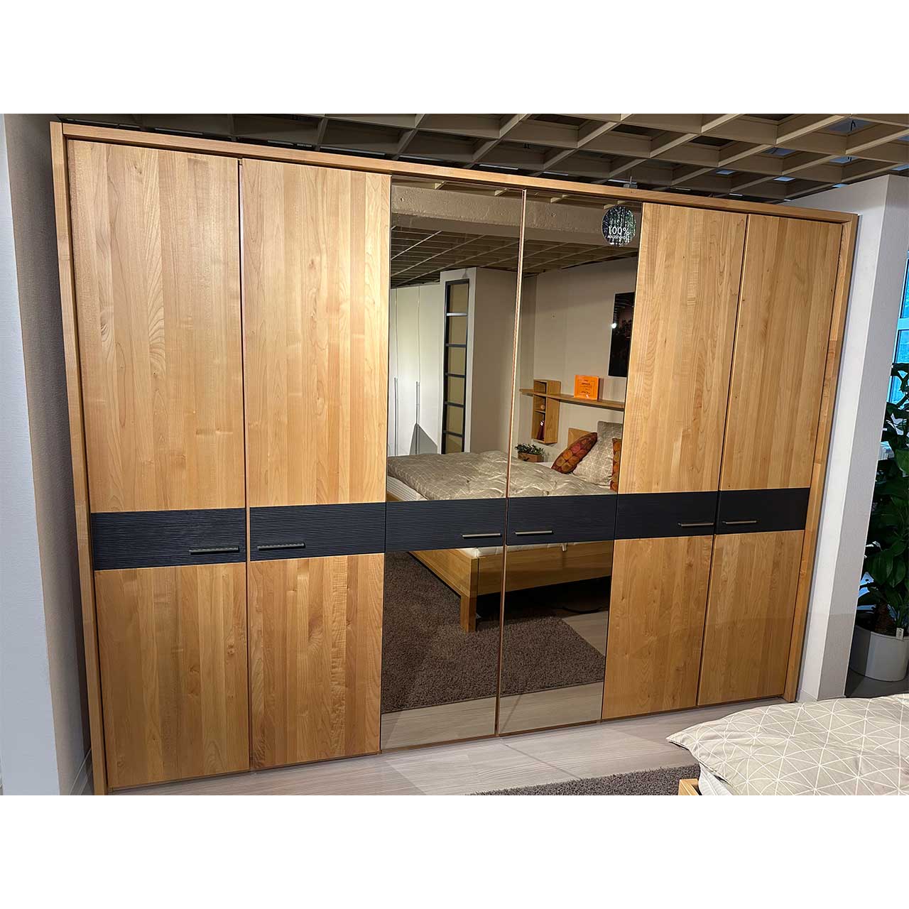 Der Kleiderschrank besteht aus rechts und links je zwei Holztüren und in der Mitte zwei Spiegeltüren. Ein grau-braunes Band läuft als Dekoration mittig durch alle Türen. Daran sind auch die Griffe angebracht. Ein Holzkranz umgibt den Kleiderschrank.