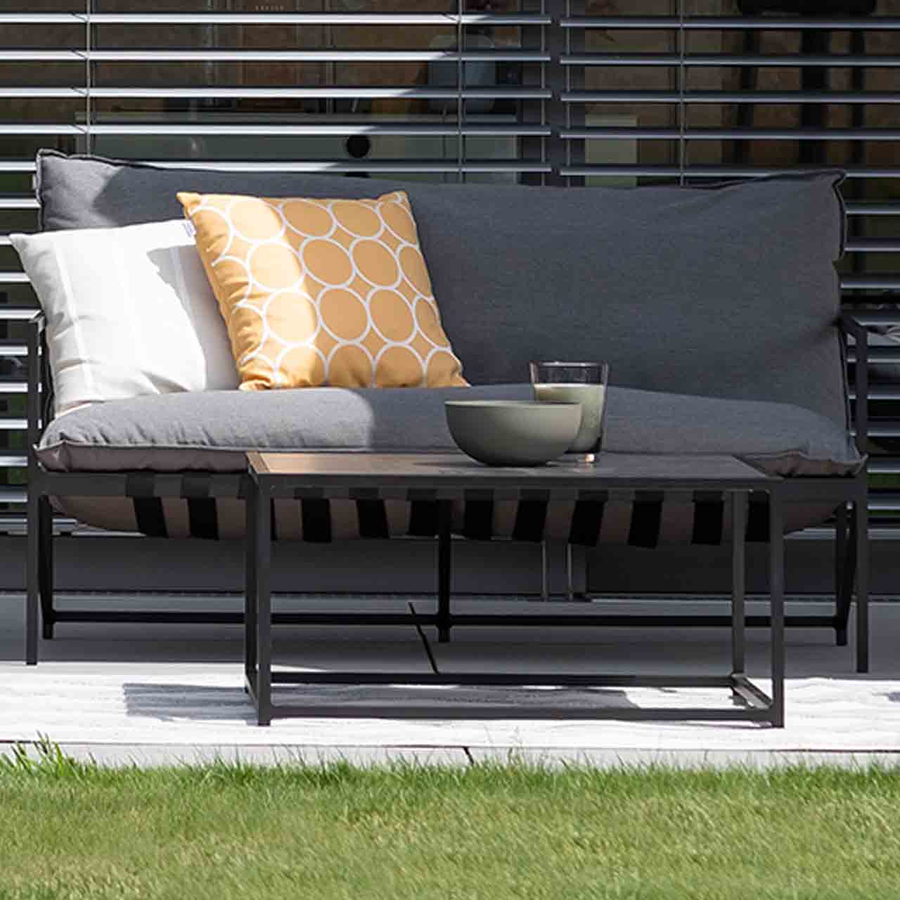 Couchtisch Ameland mit schwarzen Metallgestell und schwarzen Glas-Keramik Tischplatte steht vor dem zweisitzer Lounge Vlieland mit grauen Sitzkissen.