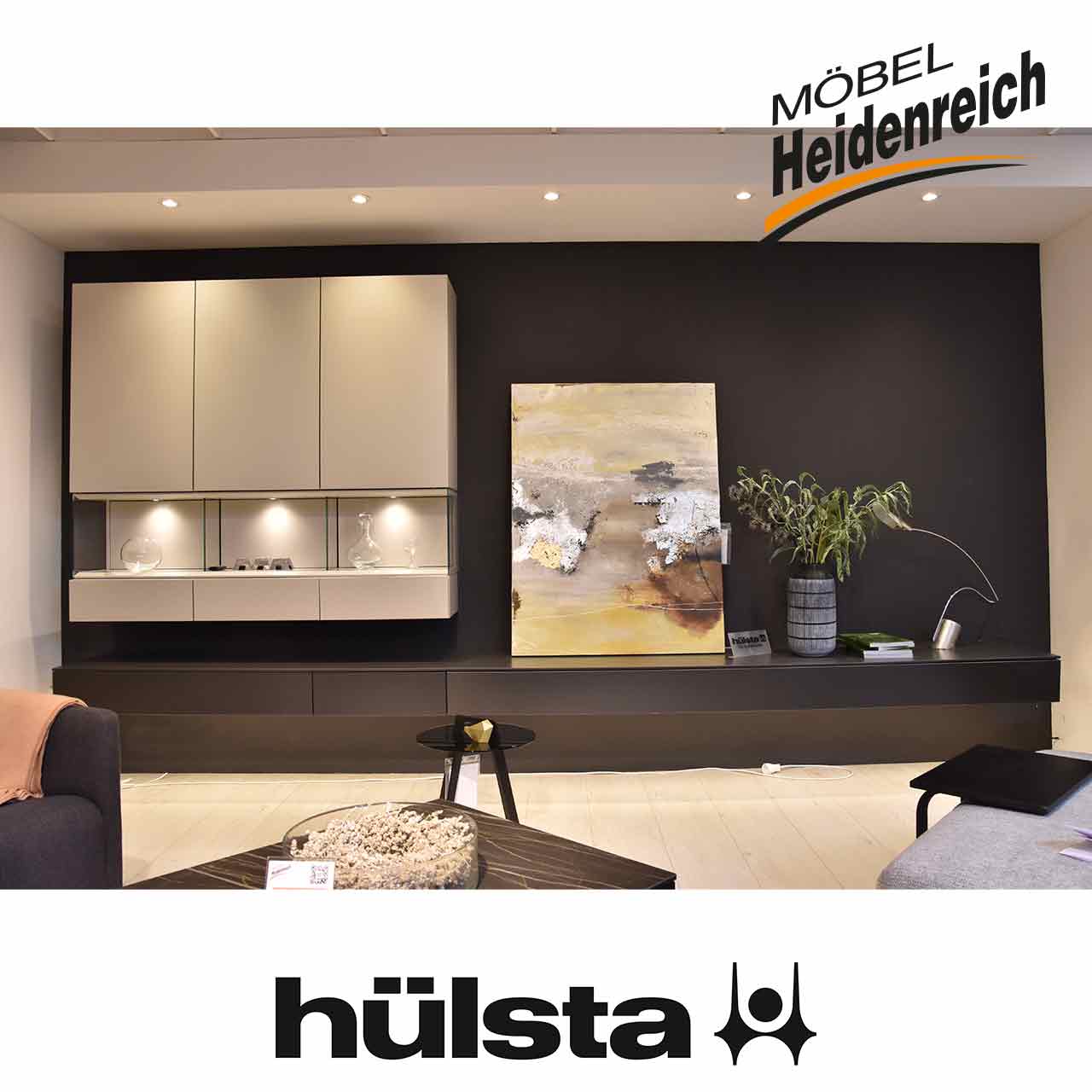 Hülsta NEO - Wohnwand seidengrau / grau Schmales dunkelgraues Lowboard Element mit zwei Schubladen und einer Klappe. Links darüber hellgraues Hängeelement mit drei Türen und drei Schubladen und Glasfach mit Beleuchtung.