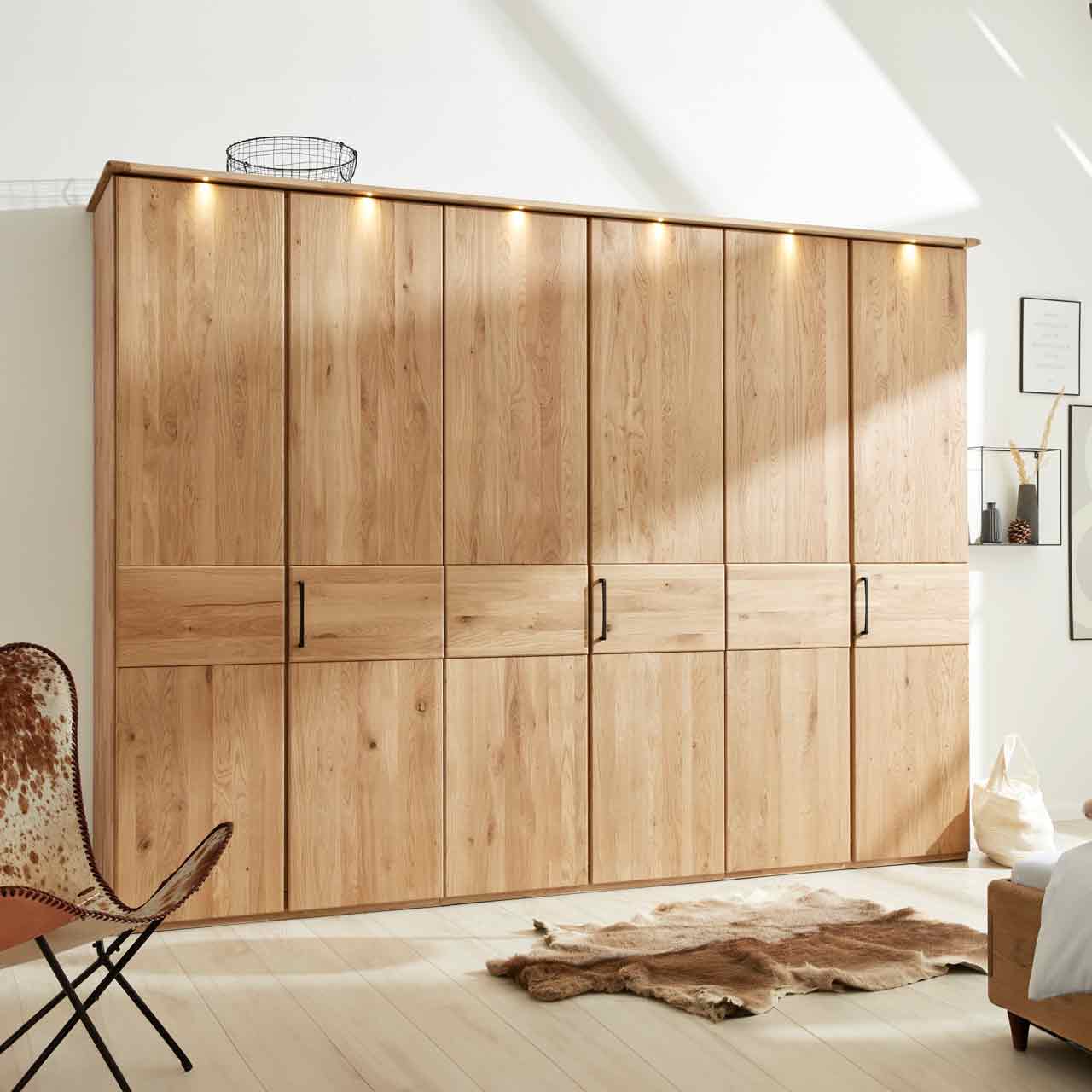 Kleiderschrank aus hellem Holz mit 6 Türen und Kranz mit Beleuchtung oben. In der Mitte eine durchlaufende optische Absetzung. An jeder zweiten Tür ist ein Griff.