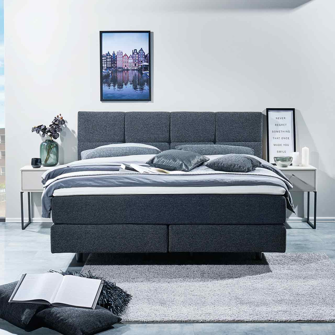 Milieuaufnahme des Boxspringbettes in einem Schlafzimmer in Frontalansicht. Das schwarze Bett besteht aus zwei Boxen unten, darüber zwei Matratzen in einem Bezug. Das Kopfteil ist durch Absteppungen in acht Quadrate unterteilt.