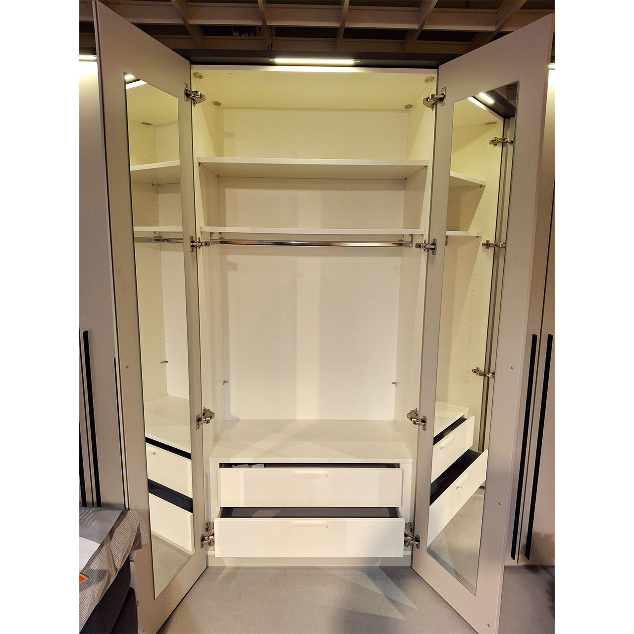 Mehrtüriger Kleiderschrank mit LED-Beleuchtung über jeder Doppeltür. Die Doppeltür in der Mitte ist geöffnet. Zu erkennen sind eine Kleiderstange, zwei Einlegeböden im oberen Drittel des Schrankes sowie zwei Schubkästen im unteren Bereich.
