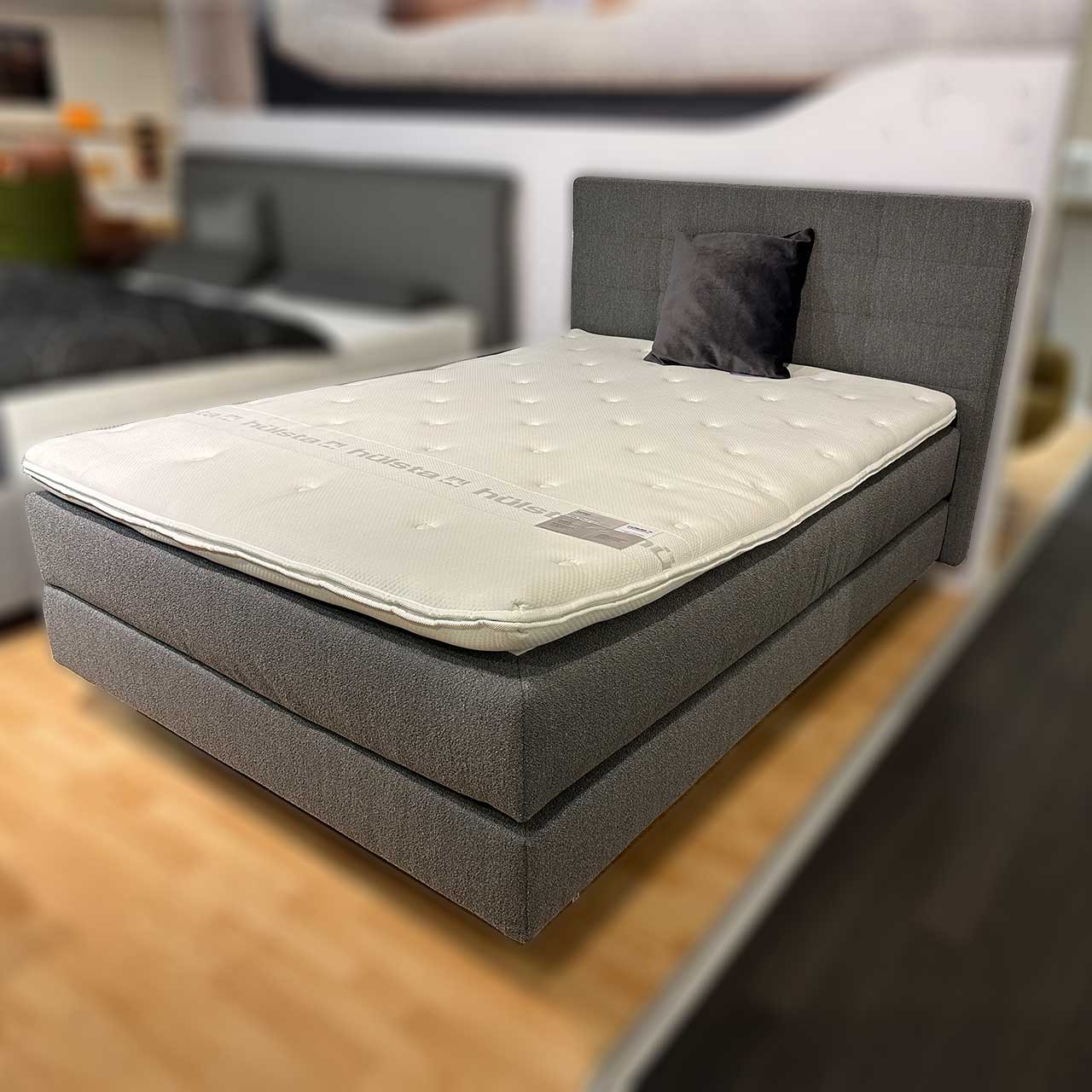 Hülsta Boxspringbett 140x200 Schmales graues Boxspringbett für eine Person im Möbelhaus fotografiert. Obenauf liegt ein unbezogener Topper mit hülsta-Logo.