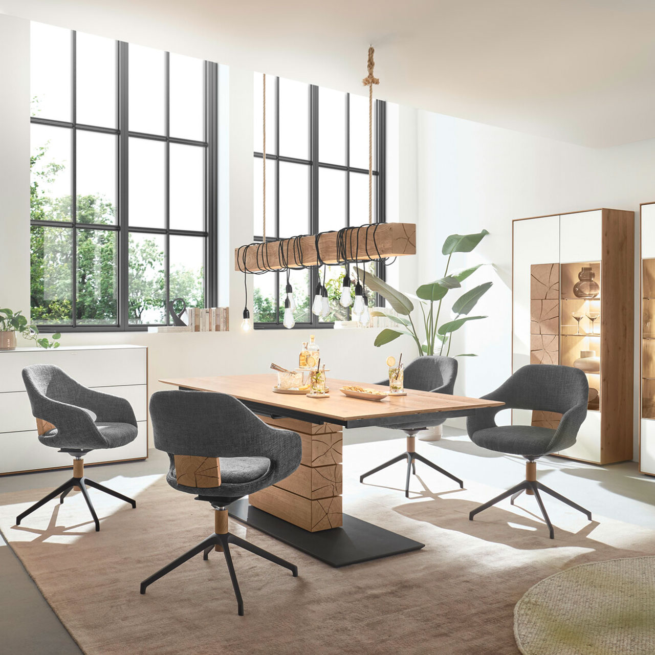MH_Kachel_Esszimmer Stilvolles Ambientebild Esszimmer mit einem hochwertigen Esstisch in Holz und anthrazitfarbenen Elementen und Spaltholzoptik im Zentralfuß, sowie vier passenden Stühlen. Im Hintergrund stehen zwei Highboards.