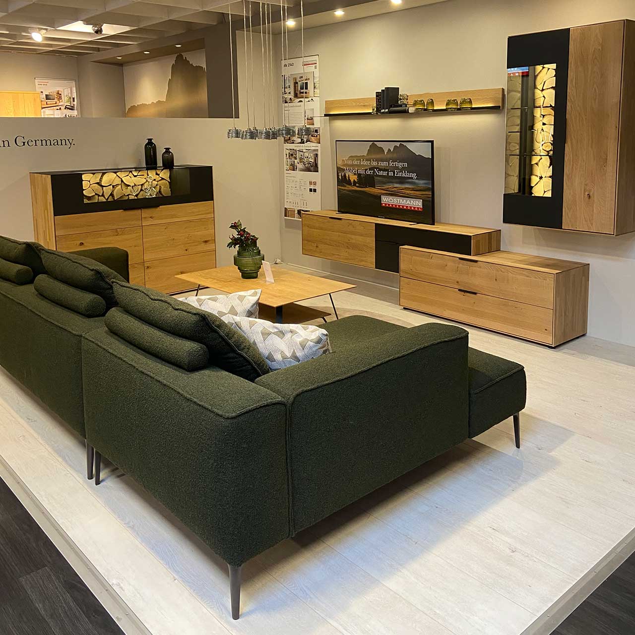 MH_Kachel_Ausstellung-Haupthaus Hier sieht man ein Bild einer Wohnzimmerlandschaft mit Sofa in dunkelgrünem Stoff 3-Sitzer, Couchtisch, Highboard mit Spaltholzakzenten, Lowboard, Hängeelement aus Massivholz.