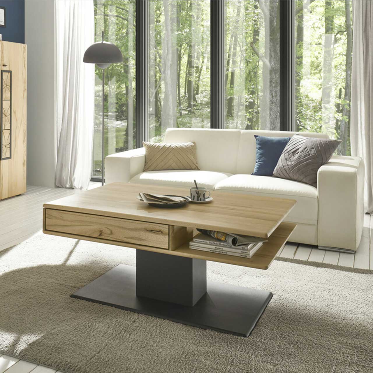 MH_Wohnzimmer_Couchtische Ambientebild mit Fokus auf einen Designer-Couchtisch mit Holzplatte und integrierter Schublade, sowie passendem Fuß aus Metall in Anthrazit und dekorativen Elementen. Im Hintergrund steht ein Sofa 2-Sitzer in Off-White.
