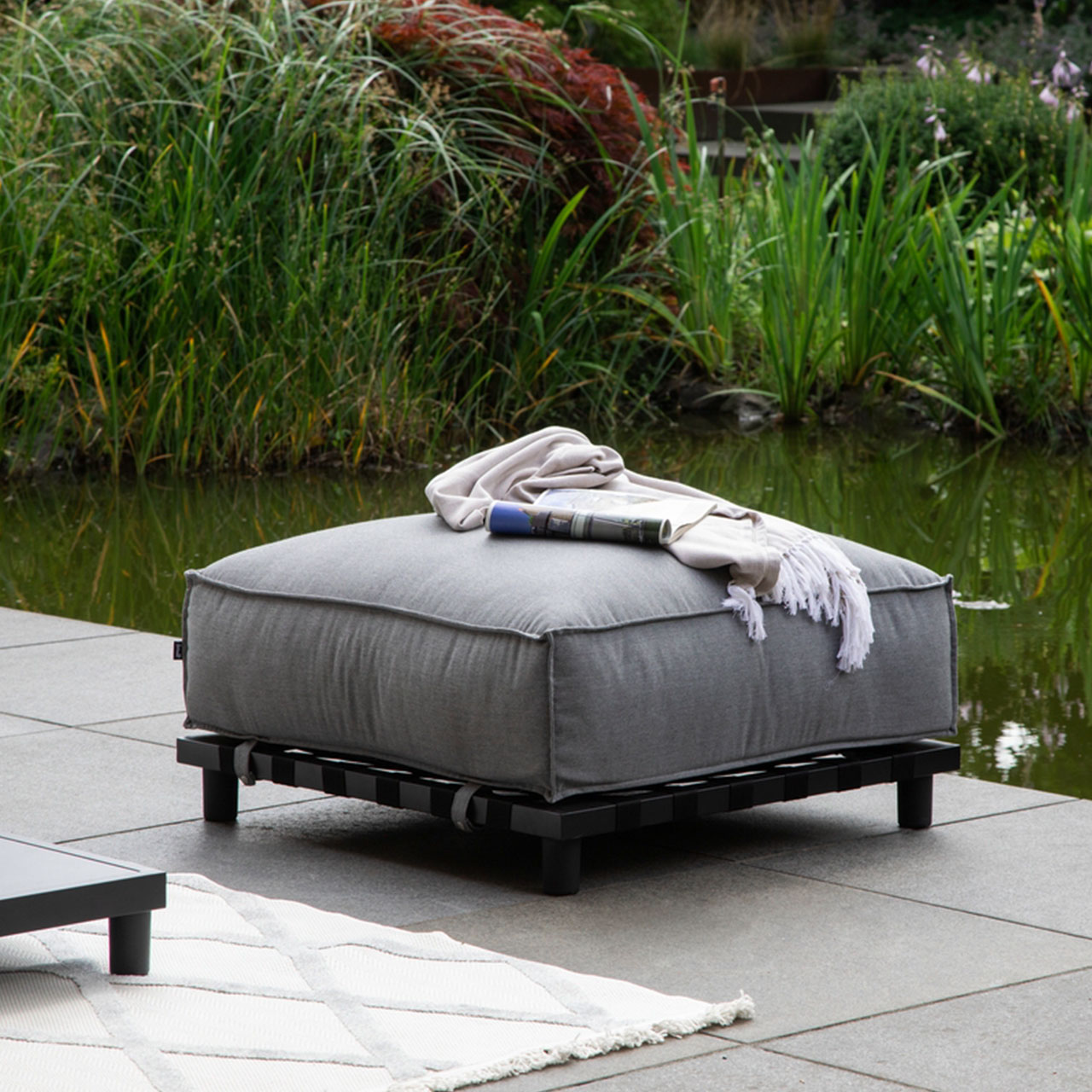 Ambiente Bild der Gartenlounge Vlieland Hocker mit schwarzen Metalluntergestell und wetterbeständigen grauen Sunbrella Stoff.