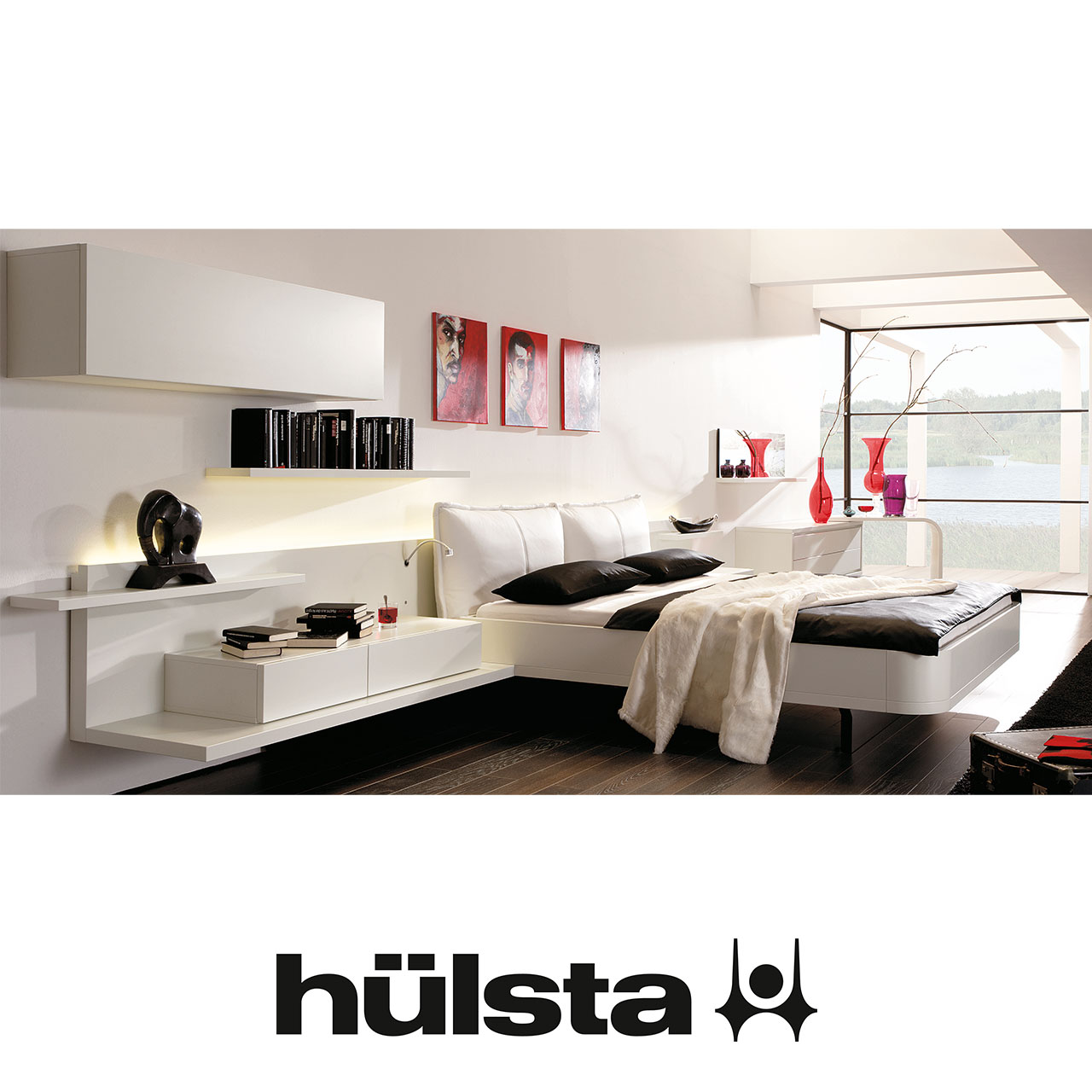Weißes Schlafzimmer. Links Hängeschrank mit griffloser Tür, darunter Wandbord und Konsole auf Podest. In der Mitte Bett mit zwei Kissenförmigen Kopfteilpolstern. Rechts Konsole und weiteres Hängeelement mit Schubladen.