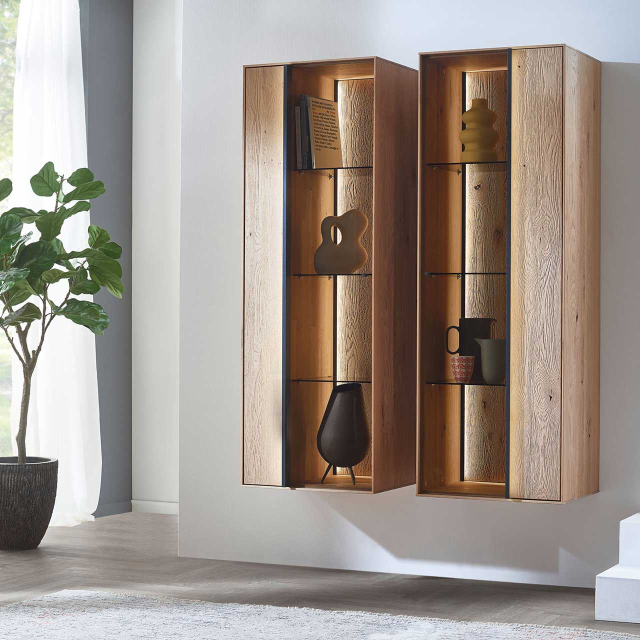 MH_Wohnzimmer_Haengeelemente Zwei passende Hängeelemente aus Holz mit schwarzen Applikationen, jeweils mit einseitig offener Struktur, indirekter Beleuchtung und dekorativen Elementen.